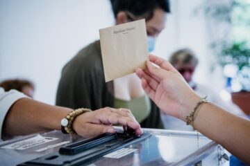 un bras montre une enveloppe au dessus d'une urne, une femme avec une motre au bras gauche ouvre l'urne à Rouvroy
