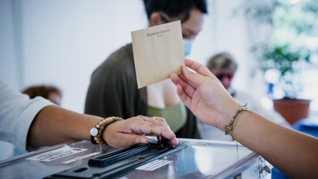 un bras montre une enveloppe au dessus d'une urne, une femme avec une motre au bras gauche ouvre l'urne à Rouvroy