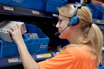 femme blonde avec les cheveux attachés avec un tee shirt orange et un casque sur les oreilles préparant une commande