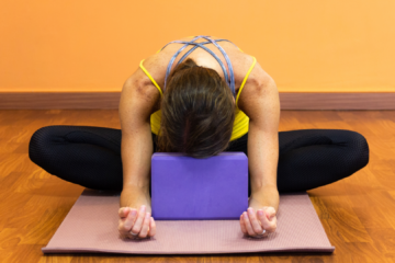 jeune fille assise en tailleur posant sa tête sur une brique de yoga bras détentus